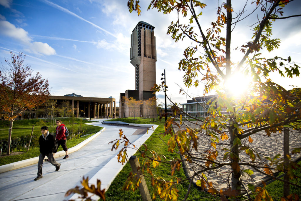 Wide-angle photo of the University of Michigan's engineering research campus with a brick tower in the center, backlit by a setting sun. Graduate students walk past the tower.